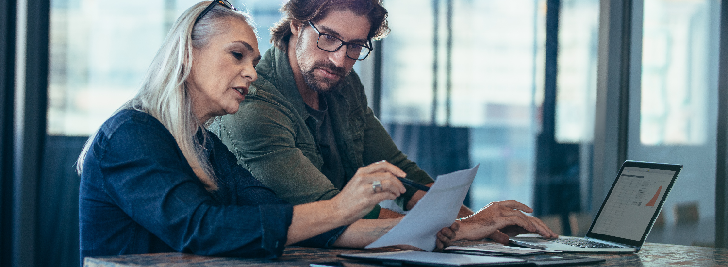 Man working on laptop while looking at a piece of paper a woman is holding and talking about